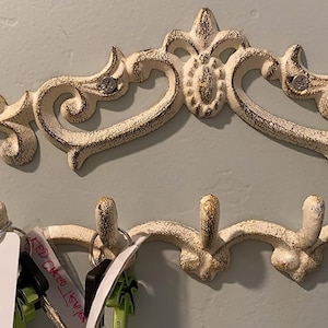 May include: White cast iron key hook with ornate scrollwork design. The hook has five hooks and is mounted on a wall. There are keys and keychains hanging on the hooks.