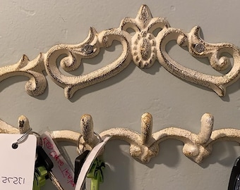 Puede incluir: Gancho para llaves de hierro fundido blanco con un diseño de volutas ornamentadas. El gancho tiene cinco ganchos y está montado en una pared. Hay llaves y llaveros colgados en los ganchos.