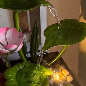 以下が含まれることがあります： ピンクの蓮の花、緑の葉、そして白い石の盆地へと流れ落ちる小さな滝のある卓上噴水。噴水は暖色系の白い光で照らされています。