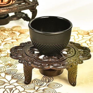 May include: A black cast iron teacup warmer with a decorative design sits on a white table with a gold floral pattern. The warmer has three legs and a round base.