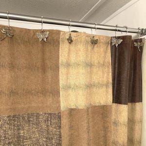 May include: A shower curtain with a patchwork design in shades of brown and tan. Decorative metal shower curtain hooks feature butterfly and dragonfly designs, adding a touch of nature to the bathroom decor.