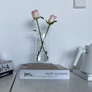 May include: A clear glass vase with a double-walled design, holding two pink and white roses. The vase sits on a stack of books, including one with the text "American Style" and a picture of the Statue of Liberty. A white kettle is visible in the background.