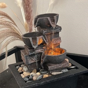 Può includere: Una piccola fontana da tavolo con un design a strati. La fontana è realizzata in materiale simile alla pietra marrone e grigia e presenta tre livelli con acqua che scende a cascata. La fontana è illuminata da luci LED bianco caldo e include pietre decorative nella base.