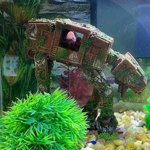 K&ouml;nnte beinhalten: Ein Aquarium mit einer braunen, moosbedeckten AT-AT Walker Dekoration. Ein rot-wei&szlig;er Fisch ist im Inneren des Walkers zu sehen. Eine leuchtend gr&uuml;ne, k&uuml;nstliche Pflanze und bunter Kies vervollst&auml;ndigen die Unterwasserumgebung.