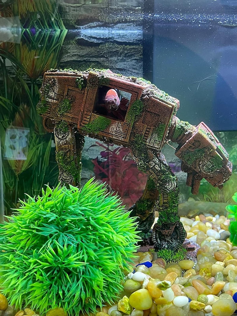 K&ouml;nnte beinhalten: Ein Aquarium mit einer braunen, moosbedeckten AT-AT Walker Dekoration. Ein rot-wei&szlig;er Fisch ist im Inneren des Walkers zu sehen. Eine leuchtend gr&uuml;ne, k&uuml;nstliche Pflanze und bunter Kies vervollst&auml;ndigen die Unterwasserumgebung.
