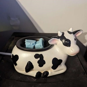 Puede incluir: Un calentador de cera de cerámica con forma de vaca blanco y negro con dos fundidos de cera azules en su interior.