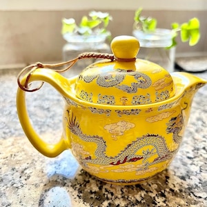 Puede incluir: Una tetera de cerámica amarilla con un diseño de dragón. La tetera tiene una tapa con una pequeña asa y un asa más grande en el lateral. La tetera está decorada con un dragón y nubes.
