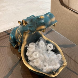 May include: A teal blue ceramic hippopotamus figurine with a gold trim. The hippo's mouth is open and is being used as a bowl to hold a large quantity of white round mints.