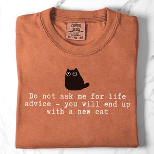 May include: A rust-colored t-shirt featuring a black cat illustration and the white text: Do not ask me for life advice - you will end up with a new cat. The shirt has a Comfort Colors brand label at the neckline indicating size XL-TG-EG.