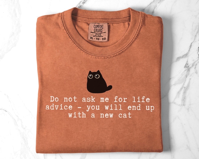 May include: A rust-colored t-shirt featuring a black cat illustration and the white text: Do not ask me for life advice - you will end up with a new cat. The shirt has a Comfort Colors brand label at the neckline indicating size XL-TG-EG.