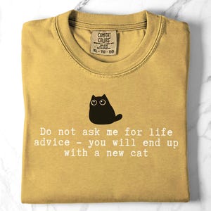 May include: A mustard yellow t-shirt featuring a black graphic of a cat and the white text: Do not ask me for life advice - you will end up with a new cat. The shirt has a crew neckline and a Comfort Colors brand label at the inner collar.