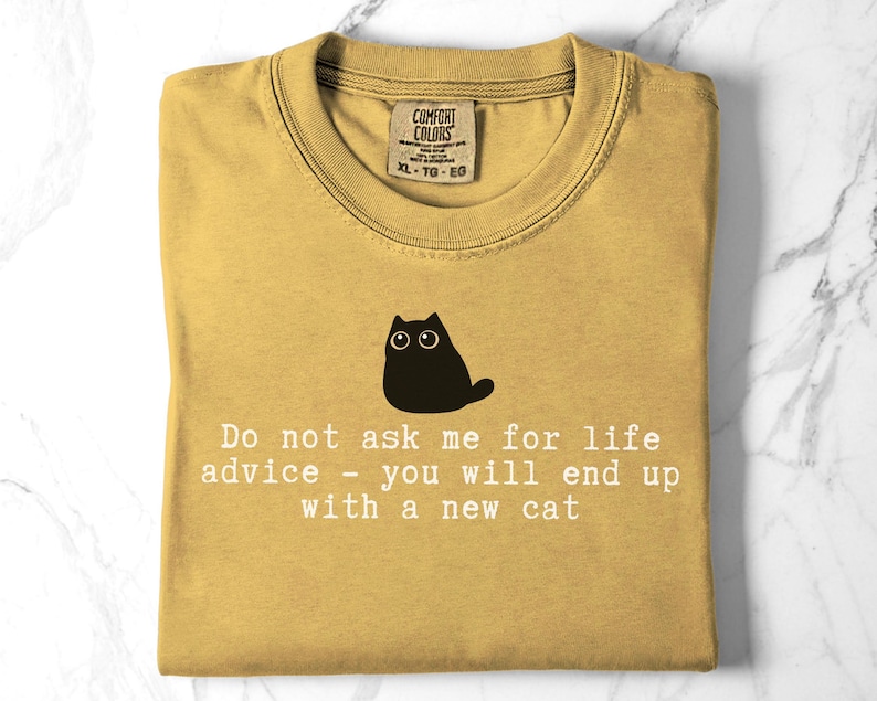 May include: A mustard yellow t-shirt featuring a black graphic of a cat and the white text: Do not ask me for life advice - you will end up with a new cat. The shirt has a crew neckline and a Comfort Colors brand label at the inner collar.