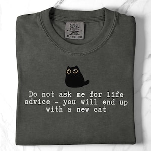 May include: A charcoal gray t-shirt featuring a graphic of a small black cat with wide eyes. Below the cat, white text reads: Do not ask me for life advice - you will end up with a new cat. The shirt has a Comfort Colors brand label at the neck.