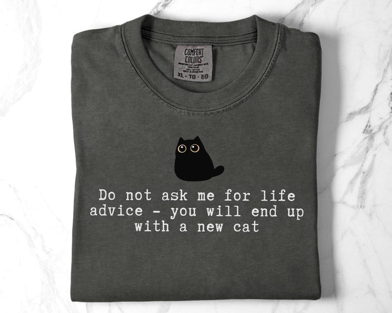 May include: A charcoal gray t-shirt featuring a graphic of a small black cat with wide eyes. Below the cat, white text reads: Do not ask me for life advice - you will end up with a new cat. The shirt has a Comfort Colors brand label at the neck.