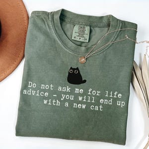 May include: An olive green short-sleeve t-shirt featuring a graphic of a small black cat with wide eyes. Below the cat, white text reads: Do not ask me for life advice - you will end up with a new cat. The shirt has a crew neckline and a tag.