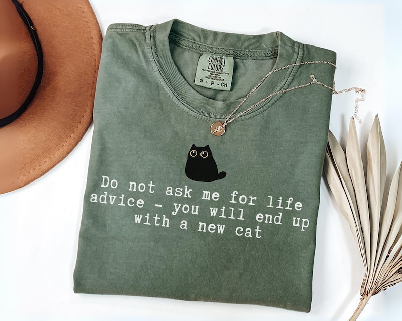 May include: An olive green short-sleeve t-shirt featuring a graphic of a small black cat with wide eyes. Below the cat, white text reads: Do not ask me for life advice - you will end up with a new cat. The shirt has a crew neckline and a tag.
