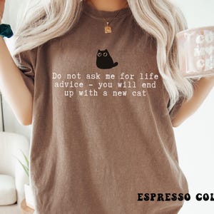 May include: An espresso-colored t-shirt featuring a graphic of a small black cat with wide eyes. Below the cat, the text reads: Do not ask me for life advice - you will end up with a new cat. The shirt has a crew neckline and short sleeves.