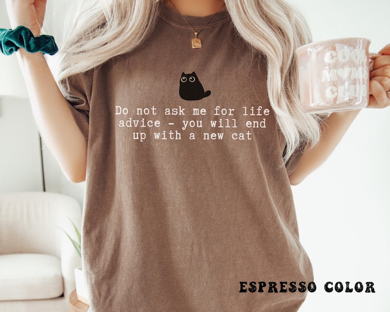 May include: An espresso-colored t-shirt featuring a graphic of a small black cat with wide eyes. Below the cat, the text reads: Do not ask me for life advice - you will end up with a new cat. The shirt has a crew neckline and short sleeves.
