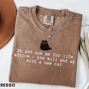 May include: An espresso-colored t-shirt featuring a black cat graphic and the white text: Do not ask me for life advice - you will end up with a new cat. The shirt has a crew neckline and a Comfort Colors brand label visible at the inner collar.