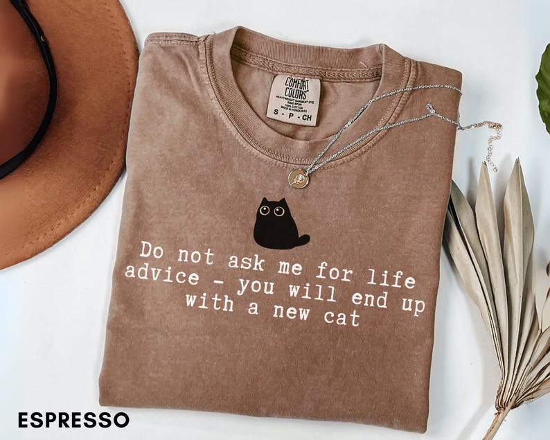 May include: An espresso-colored t-shirt featuring a black cat graphic and the white text: Do not ask me for life advice - you will end up with a new cat. The shirt has a crew neckline and a Comfort Colors brand label visible at the inner collar.