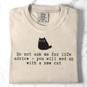 May include: A beige t-shirt featuring a black graphic of a cat and the text: Do not ask me for life advice - you will end up with a new cat. The shirt has a Comfort Colors brand label at the neckline indicating size XL, TG, EG, and 100% cotton material.