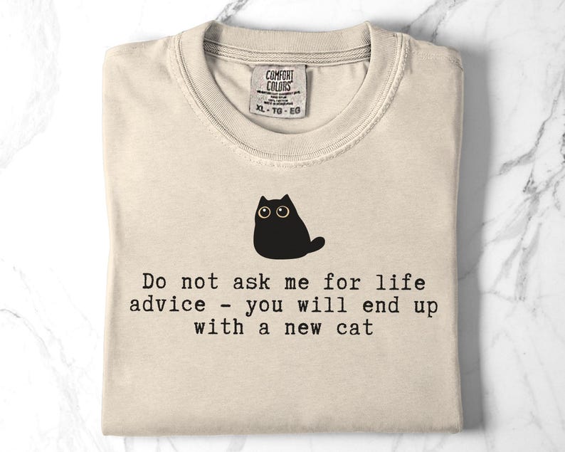May include: A beige t-shirt featuring a black graphic of a cat and the text: Do not ask me for life advice - you will end up with a new cat. The shirt has a Comfort Colors brand label at the neckline indicating size XL, TG, EG, and 100% cotton material.