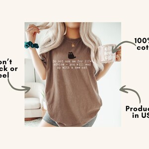 May include: A brown short-sleeve t-shirt featuring a small black cat graphic and the white text: Do not ask me for life advice - you will end up with a new cat. The shirt is made of 100% US cotton and is produced in the USA with a durable print.