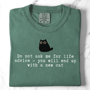 May include: A sage green t-shirt featuring a black cat illustration and the white text: Do not ask me for life advice - you will end up with a new cat. The shirt has a Comfort Colors brand label at the inner neckline, indicating size XL-TG-EG.