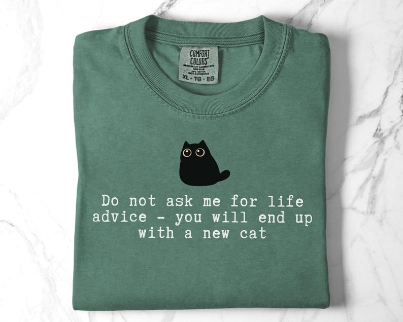 May include: A sage green t-shirt featuring a black cat illustration and the white text: Do not ask me for life advice - you will end up with a new cat. The shirt has a Comfort Colors brand label at the inner neckline, indicating size XL-TG-EG.
