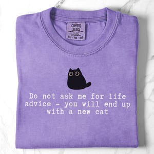 May include: A purple t-shirt featuring a graphic of a black cat with large eyes. Below the cat, white text reads: Do not ask me for life advice - you will end up with a new cat. The shirt has a Comfort Colors brand label at the inner neckline.