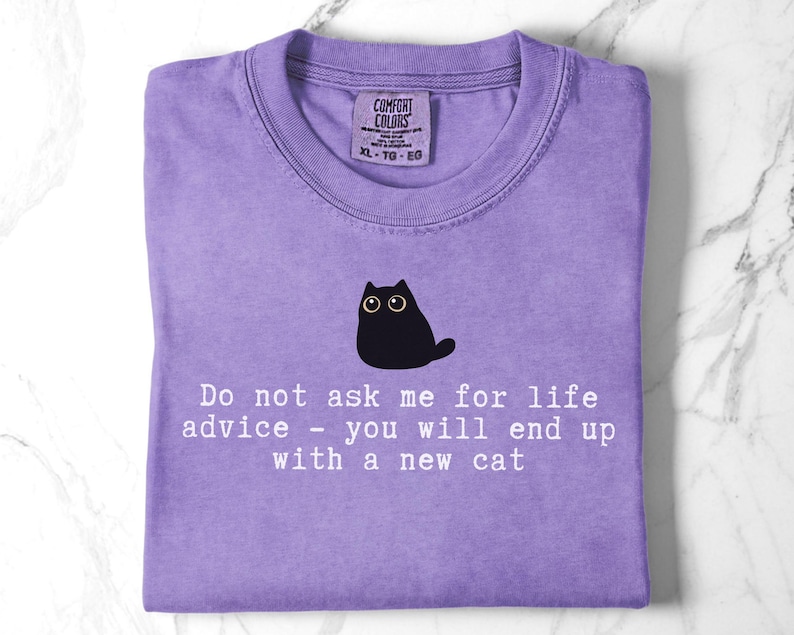 May include: A purple t-shirt featuring a graphic of a black cat with large eyes. Below the cat, white text reads: Do not ask me for life advice - you will end up with a new cat. The shirt has a Comfort Colors brand label at the inner neckline.