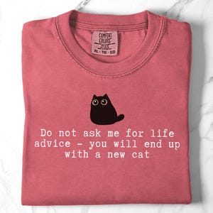 May include: A dusty rose crewneck t-shirt featuring a black graphic of a wide-eyed cat above the white text: Do not ask me for life advice - you will end up with a new cat. The shirt has a Comfort Colors label at the inner neckline showing size XL.