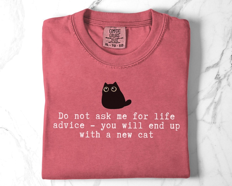 May include: A dusty rose crewneck t-shirt featuring a black graphic of a wide-eyed cat above the white text: Do not ask me for life advice - you will end up with a new cat. The shirt has a Comfort Colors label at the inner neckline showing size XL.