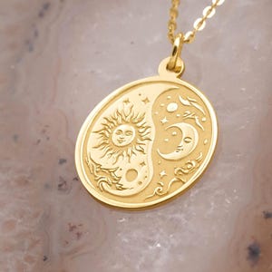 May include: A gold-colored pendant necklace featuring an oval pendant with sun and moon designs. The pendant is suspended from a delicate chain. The necklace is displayed on a light-colored, patterned surface.