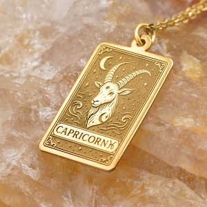 Puede incluir: Un collar con colgante rectangular dorado con la palabra "CAPRICORN" en la parte inferior. El colgante presenta una ilustración de cabra con una luna creciente y estrellas. El collar se muestra sobre una piedra marrón claro.