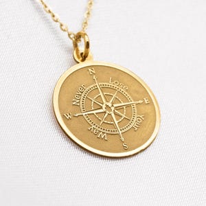 Puede incluir: Un collar con colgante dorado con un diseño de brújula circular. El colgante tiene la inscripción "Never Lose Your Way". La cadena del collar es fina y dorada.