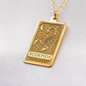 Puede incluir: Un collar con colgante dorado con un colgante rectangular con un diseño detallado de escorpión, una luna creciente y la palabra "SCORPIO". La cadena del collar también es dorada. El colgante es probablemente de metal dorado.