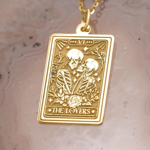 Pode incluir: Colar com pingente dourado com um berloque retangular com um desenho de carta de tarô. O berloque retrata dois esqueletos se abraçando, com as palavras "THE LOVERS" na parte inferior. O colar está em uma corrente dourada.