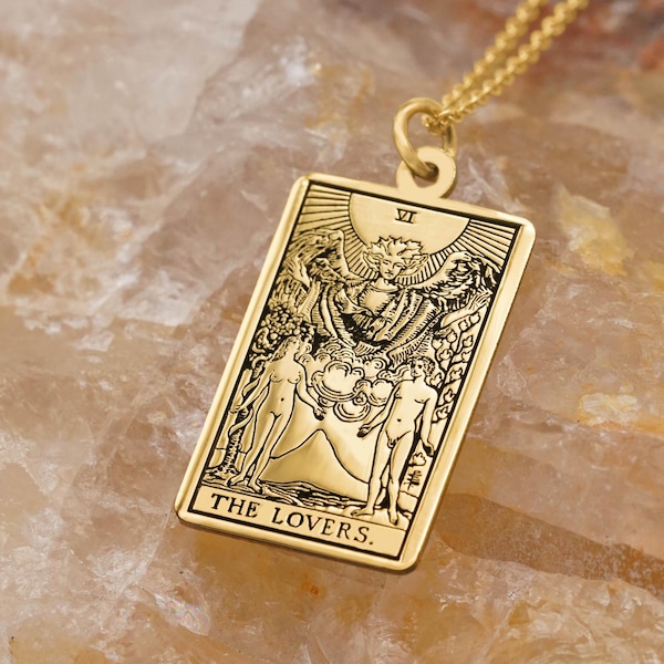 Pode incluir: Colar com pingente retangular dourado com o design da carta de tarô dos Amantes. O pingente tem detalhes gravados intrincados de figuras e símbolos. A corrente é delicada e dourada. O pingente é exibido em uma superfície clara e texturizada.