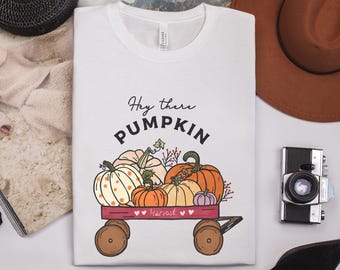 Puede incluir: Camiseta blanca con un diseño de calabazas en un carro rojo, con el texto "Hey there Pumpkin" y "Harvest". Las calabazas son naranjas, blancas y rosas. El carro tiene ruedas marrones.
