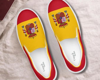 Puede incluir: Un par de zapatillas sin cordones con el diseño de la bandera nacional de España, con un centro amarillo y bandas rojas en la puntera y el talón. El escudo de armas de España está impreso en el centro. La plantilla interior muestra el texto Unpack Relax.