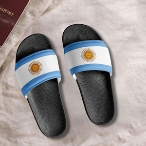 Puede incluir: Un par de sandalias deslizantes negras con el diseño de la bandera argentina. Las sandalias tienen una banda blanca con un borde azul y un emblema de sol dorado. Las sandalias están sobre una superficie blanca.