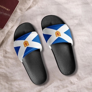 May include: Pair of black slide sandals featuring the Nova Scotia flag design. The sandals have a blue and white flag pattern across the top with a gold emblem. The sandals are shown on a light beige surface.