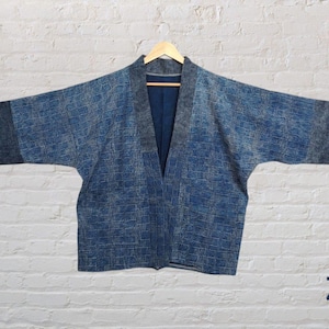 May include: A blue denim kimono with a textured pattern. The kimono has a V-neck and long sleeves. The kimono is hanging on a wooden hanger against a white brick wall. Z2