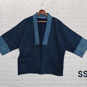 Op de afbeelding: Een marineblauwe jas in kimono-stijl met een contrasterende lichtblauwe kraag en manchetten. De jas is gemaakt van een textuurstof en hangt aan een houten hanger tegen een witte bakstenen muur. De letters "SS" zijn zichtbaar.