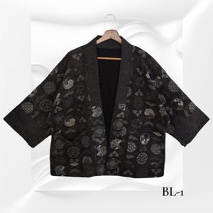 Può includere: Giacca grigio scuro in stile kimono con interno nero, appesa a una gruccia di legno. La giacca presenta un motivo ripetuto di disegni circolari e geometrici bianchi e grigi. Il testo "BL-1" è visibile nell'angolo in basso a destra.