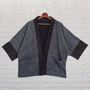 Puede incluir: Chaqueta gris estilo kimono con ribetes y bolsillos negros, colgada de una percha de madera. La chaqueta tiene mangas anchas y un aspecto texturizado, ofreciendo un look casual pero elegante. El fondo es una pared de ladrillos blancos.