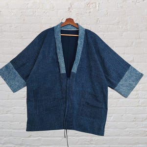Puede incluir: Chaqueta azul estilo kimono con un patrón de rayas. La chaqueta presenta un cuello y puños azul claro contrastantes. Cuelga de una percha de madera contra una pared de ladrillos blancos.