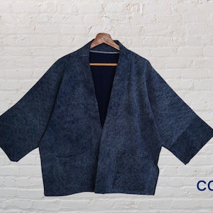 May include: A dark blue indigo-dyed jacket with a textured, speckled pattern, wide sleeves, and an open front design. The garment features two front pockets and is displayed on a wooden hanger. The text CC-1 is visible in the bottom right corner.