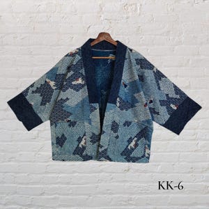 Könnte beinhalten: Blau-weiß gemusterte Jacke im Kimono-Stil mit dunkelblauem Kragen und Manschetten. Die Jacke hat ein Wellen- und Vogelmuster. Das Kleidungsstück hängt an einem Holzbügel vor einer weißen Backsteinmauer. Der Text "KK-6" ist sichtbar.